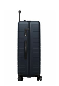 Valise rigide de couleur bleu marine foncé, avec des rainures verticales, une serrure TSA, une poignée rétractable et quatre roulettes pivotantes noires pour la mobilité.