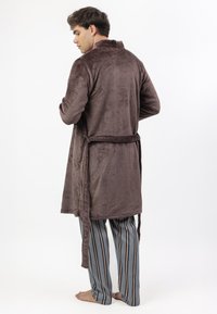 Hombre vestido con una bata de felpa marrón con cinturón y pantalones de pijama a rayas azules, de pie descalzo frente a un fondo blanco liso.