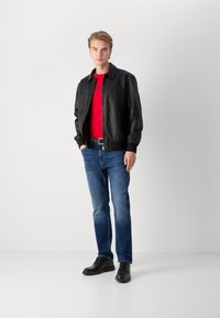 Črna usnjena bomberska jakna čez rdečo majico, modri kavbojki in črnimi vezalkami. Model stoji proti nevtralni ozadju.