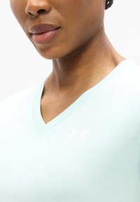 Ανοιχτό μπλε-πράσινο T-shirt με V λαιμόκοψη, φτιαγμένο από απαλό ύφασμα, με διακριτικό λευκό λογότυπο Under Armour στο αριστερό στήθος, κοντινή προβολή της λαιμόκοψης.
