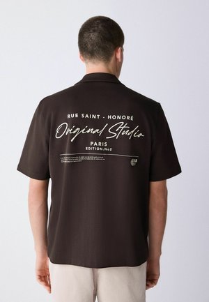 Kurzärmeliges Hemd mit Kragen aus dunkelbraunem Stoff, auf dessen Rückseite weißer Drucktext zu sehen ist, darunter "Rue Saint-Honoré" und "Original Studio".