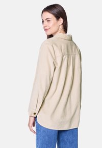 Beige corduroy shirt with a relaxed fit, button cuffs, and a back yoke detail. Smooth texture and rounded hemline, paired with blue jeans.
