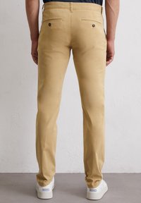 Beige katoenen chino's met achterzakken, voorzien van twee knopen. Gestoorde pijpvorm en een gladde, matte textuur. Draagt met witte sneakers.
