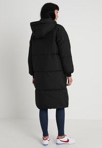 Doudoune noire avec capuche, design long, texture matelassée et poignets côtelés, portée sur un jean bleu et des sneakers blanches avec des accents rouges.