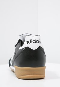 Zwarte leren sneaker met witte accenten, voorzien van een gevoerde kraag en een gumrubberen zool. Adidas-logo op de hak.