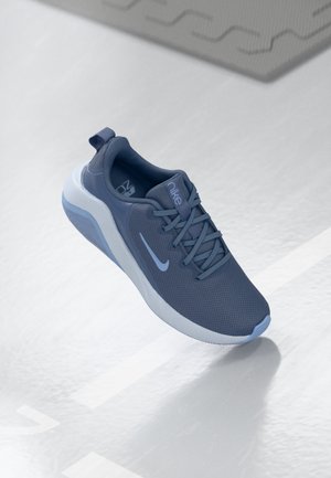 Mørkeblå Nike løbesko med hvid sål, der svæver over en lyst grå tekstureret gulv med geometriske mønstre.