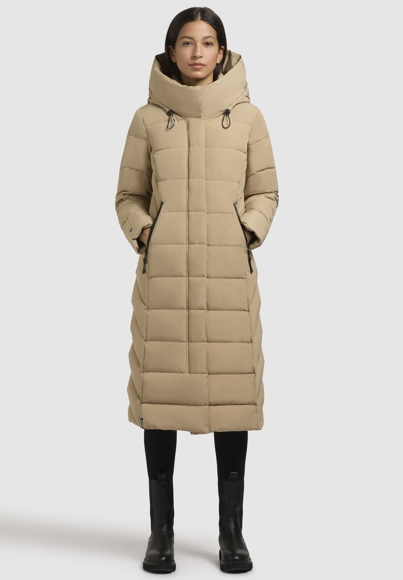 Nainen, jolla on pitkä beige palttinavuorattu takki, jossa on korkea kaulus ja kädet vetoketjullisissa taskuissa, yhdistettynä mustiin leggings-housuihin ja saappaisiin.