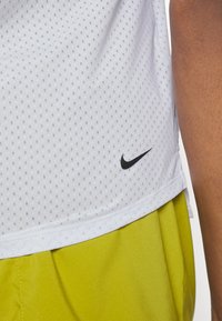 Witte, textuurvolle, kortebloes sportshirt met kleine perforaties; met een zwart Nike-logo aan de onderkant links. Gecombineerd met felgele shorts.