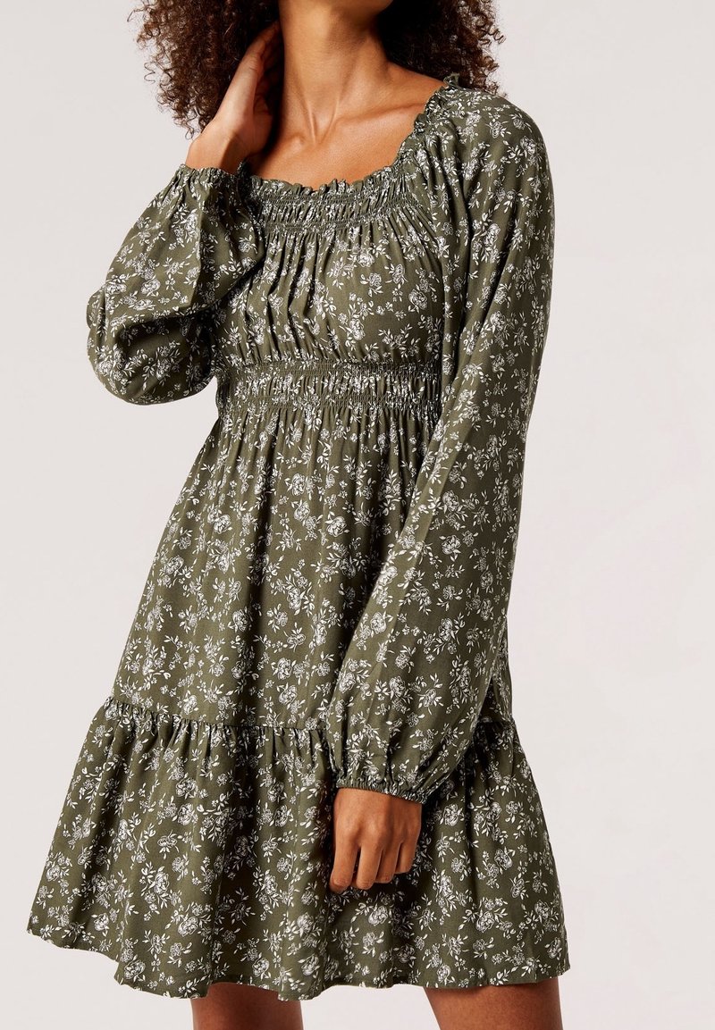 Femme portant une robe fleurie vert olive à manches longues, corsage smocké et ourlet volanté, posant avec une main près du cou.