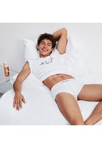 Un t-shirt et un slip blancs Calvin Klein, avec une texture lisse, portés par une personne allongée sur des draps blancs ; à côté se trouve un produit CK dans une bouteille rose.