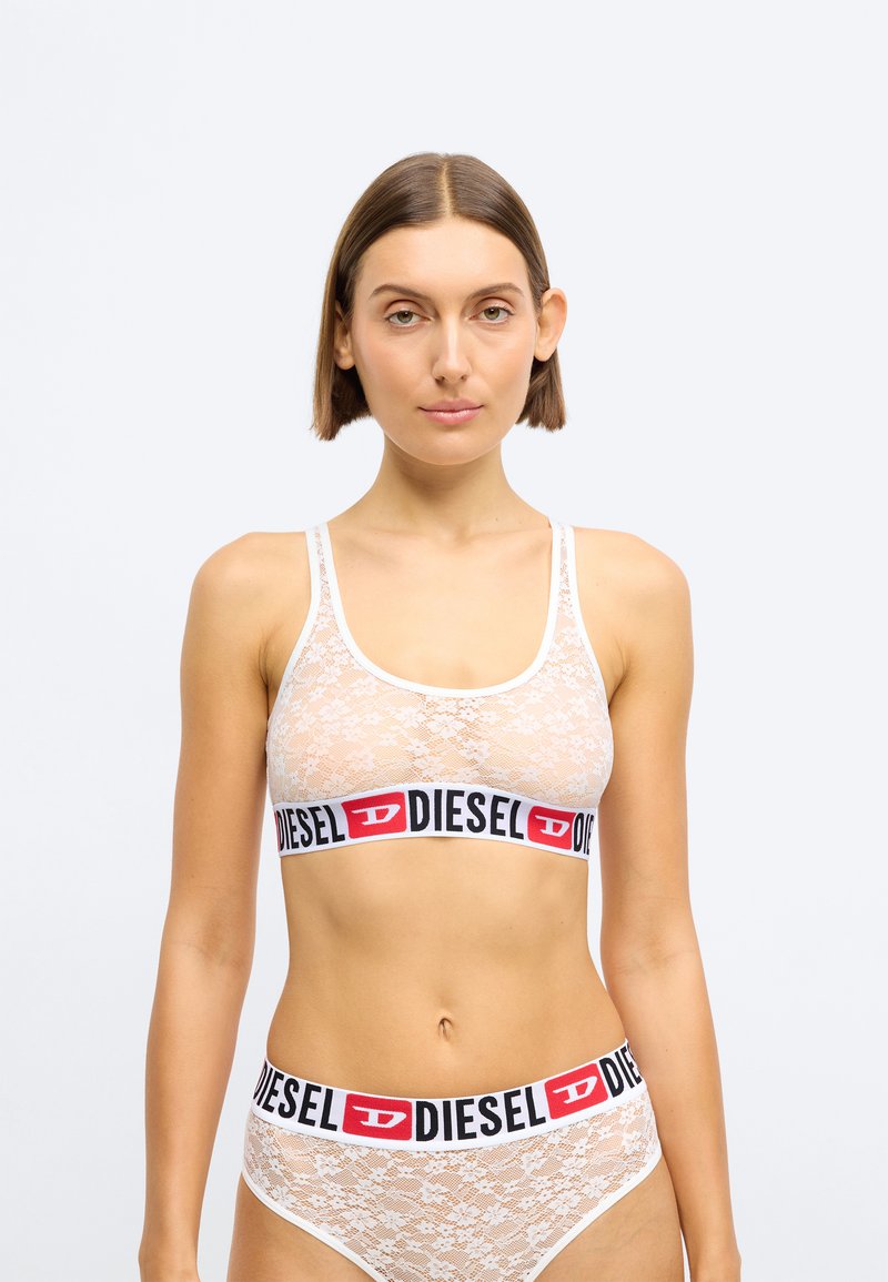 Brassière en dentelle blanche avec une large bande logo Diesel noire et rouge, ornée de motifs floraux délicats et de bretelles réglables. Bikini en dentelle assorti présenté.