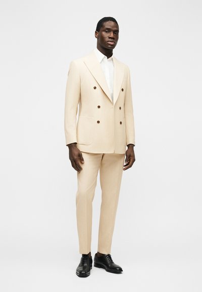 Άνδρας που φοράει ανοιχτό μπεζ, διπλό σακάκι με καφέ κουμπιά, λευκό πουκάμισο και μαύρα παπούτσια, στέκεται μπροστά σε μια απλή φόντο.