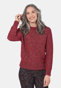Rood, gestructureerd brei-sweater met een ronde halslijn, geribbelde boorden en zoom. Model draagt een patroon met donkere broek; minimaal achtergrond.