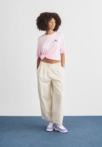 Light pink knotted t-shirt with logo, cream wide-leg trousers, and purple-accented sneakers, set against a minimalist background.