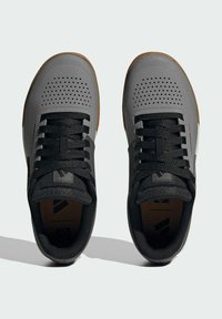 Šedé sportovní boty s černými tkaničkami, perforovaný design, polstrovaný límec a hnědá gumová podrážka. Obsahují branding na jazyku a straně.