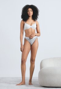 Fehér melltartó sima anyaggal és állítható pántokkal, szürke, derékpántos női alsóneművel párosítva. A modell semleges színű szőnyegen áll.