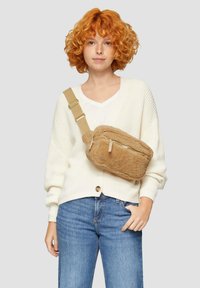 Beige Crossbody-Tasche mit Reißverschluss, verstellbarem Tragegurt und zwei Quaste-Details an der Vorderseite. Getragen über einem weißen Oberteil und Jeans.