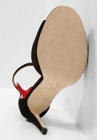 Czarne zamszowe sandały na wysokim obcasie z paskiem wokół kostki, odkryty palec, czerwone wnętrze i gładka beżowa podeszwa, ustawione pod kątem w białym rogu.