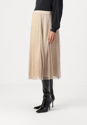 Vrouw draagt een beige geplooide midi-rok, zwarte top met lange mouwen en zwarte hoge hak laarzen met puntige neus, staand zijwaarts.