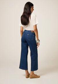 Jeans anchos de color azul con cintura alta, que presentan bolsillos traseros, una etiqueta de cuero marrón y una textura de denim suave. Se combinan con zapatos de color marrón claro.