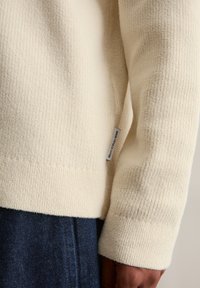 Sweter w kolorze kremowym o fakturowej strukturze z prążkowanym wzorem. Posiada metkę z boku i jest zestawiony z ciemnoniebieskimi dżinsami.