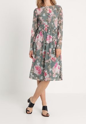 Vrouw draagt een knielange groene jurk met roze en witte bloemenprint en lange mouwen, gecombineerd met zwarte open-toe sandalen met blokhak.