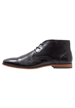 Chaussure habillée noire en cuir pour cheville avec fermeture à lacets, bout pointu et semelle marron empilée sur fond blanc.