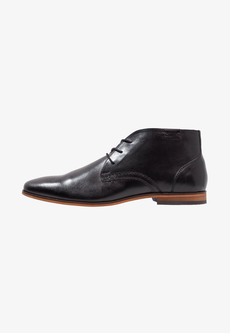 Chaussure habillée noire en cuir pour cheville avec fermeture à lacets, bout pointu et semelle marron empilée sur fond blanc.