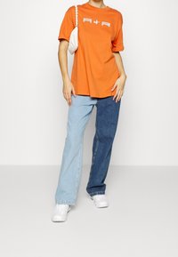 Oranžové oversized tričko s bílým logem, doplněné barevně blokovanými džínami (světle modré a tmavě modré). Bílé tenisky a malá taška.