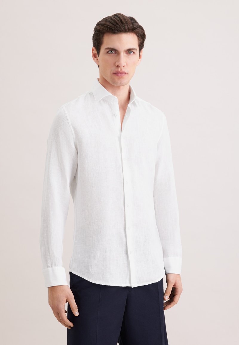 Homme aux cheveux noirs portant une chemise blanche à manches longues à boutons et un pantalon sombre, debout devant un fond clair uni.