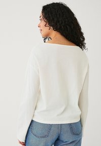 Witte lange mouw top met een brede halslijn en een zachte, gladde textuur, gecombineerd met blauwe jeans. Het zicht toont de achterkant van het shirt.