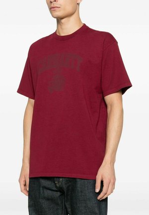 Giovane uomo che indossa una t-shirt bordeaux tinta unita a maniche corte e jeans blu scuro, in piedi davanti a uno sfondo bianco.