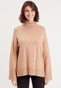 Pull à col roulé beige avec une coupe décontractée, des poignets et un ourlet côtelés, et un tissu doux et texturé. Le mannequin le porte avec un pantalon sombre.