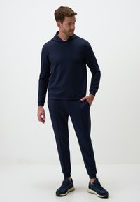 Felpa navy con cappuccio, maniche lunghe e polsini a costine. Abbinata a pantaloni tapered coordinati con tasche laterali e polsini elastici.