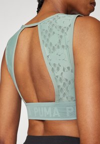 Mėtų žalios spalvos sportinis liemenėlė su nėriniu ir atvira nugara, platus dirželis su logotipo detalėmis apačioje. Lygi tekstūra.