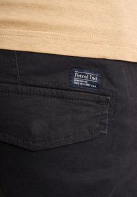 Mørkeblå jeans med tekstureret overflade, med en baglomme og en marineblå label med "Petrol Ind" og andre detaljer. Lysfarvet t-shirt ovenfor.