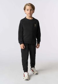 Completo felpa e pantaloni sportivi neri, caratterizzato da una texture morbida, polsini a costine e un piccolo logo ricamato sul petto. Le scarpe hanno un design beige e rosso.