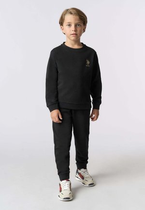 Completo felpa e pantaloni sportivi neri, caratterizzato da una texture morbida, polsini a costine e un piccolo logo ricamato sul petto. Le scarpe hanno un design beige e rosso.