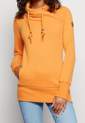 Woman wearing a bright orange hoodie with drawstring knots, front pocket, and light blue jeans, standing with one hand in pocket.