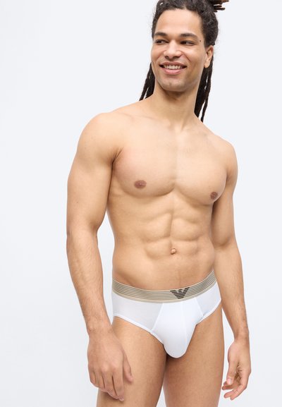 Χαμογελαστός άνδρας χωρίς πουκάμισο με ντραντλοκς, φορώντας λευκά εσώρουχα με μπεζ ριγέ μέση, σε απλό ανοιχτόφωτο φόντο.