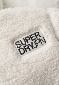 Tissu en polaire blanc avec une texture duveteuse ; l'étiquette cousue indique "SUPER DRYJPN" en noir sur un fond gris clair.