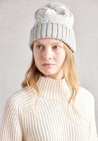 Bonnet en tricot gris avec un design côtelé et un pompon duveteux sur le dessus, associé à un pull col roulé côtelé de couleur crème.