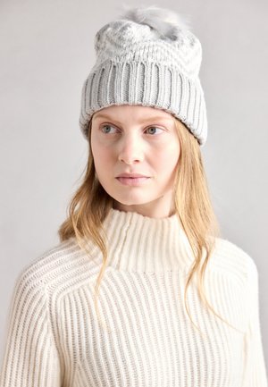 Bonnet en tricot gris avec un design côtelé et un pompon duveteux sur le dessus, associé à un pull col roulé côtelé de couleur crème.