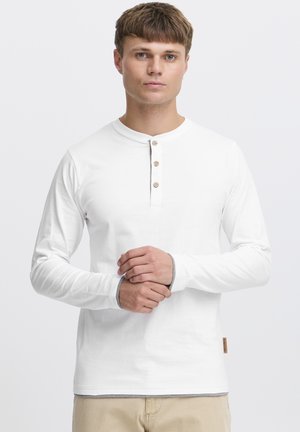 Camisa henley blanca de manga larga con cuatro botones de madera, puños grises en contraste y una sutil textura. Etiqueta en la parte inferior izquierda.