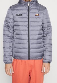 Mand iført en grå quilted zip-jakke med ellesse-logoer og lysorange bukser, stående med armene afslappet langs siden.