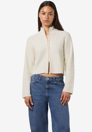 Cropped, ribbed, cream-colored sweater with a high collar and zip closure. Paired with high-waisted, wide-leg, blue denim jeans.