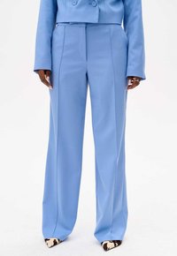 Lichtblauwe op maat gemaakte wijde pantalon met scherpe plooien aan de voorkant, gecombineerd met gemusterde hoge hakken met een koeienprint.