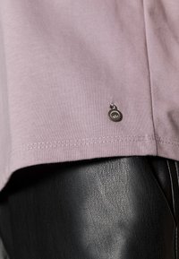 Camiseta de algodón lavanda con escote redondeado, mangas cortas y un pequeño logo metálico en el dobladillo; combinada con pantalones negros texturizados.