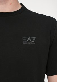 Juoda trumpomis rankovėmis medvilninė marškinėliai, pagaminti iš minkštos medvilnės, su subtiliu pilkos spalvos EA7 Emporio Armani logotipu ant kairės krūtinės.