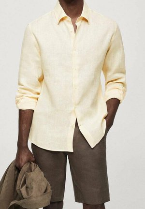 Man draagt een lichtgeel linnen overhemd met lange mouwen opgerold, bruine korte broek, houdt een bruine jas vast in de linkerhand.